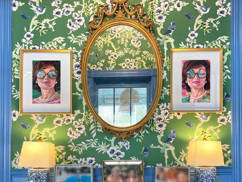 Framed 'Jackie in Palm Beach' prints displayed on floral wallpaper flanking a gold mirror, styled for the interior design trade